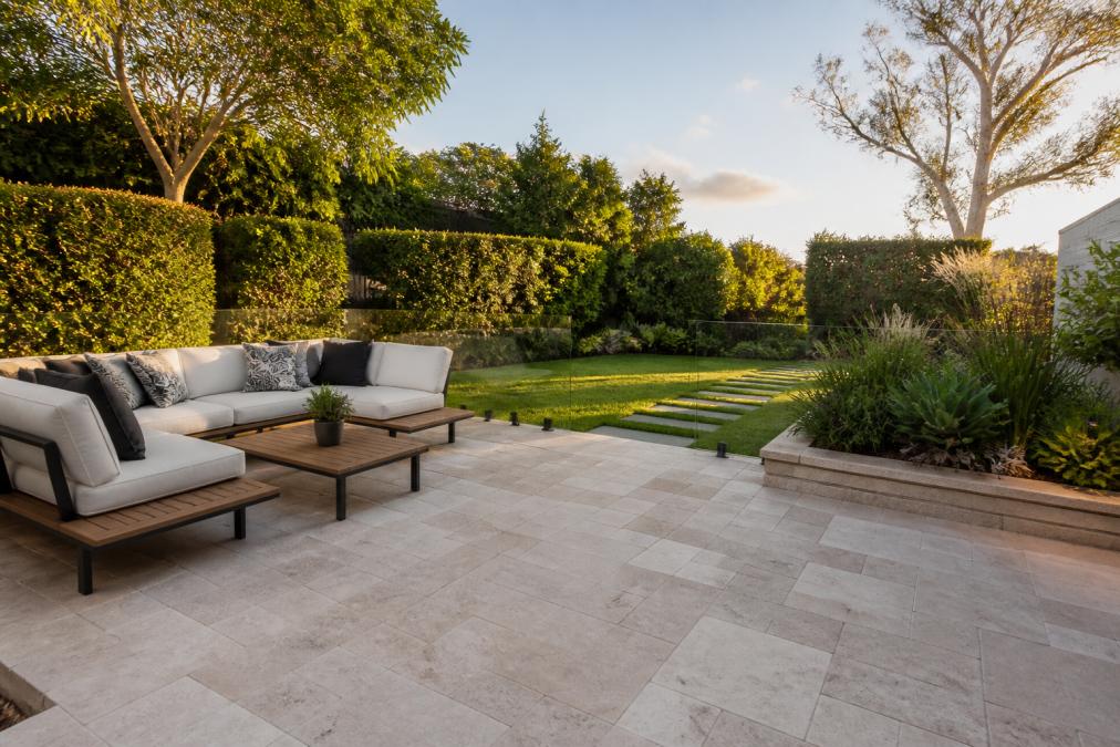 Wide residential backyard showing modern stone paver patio with warm afternoon sunlight, comfortable outdoor sectional sofa positioned on the pavers, native plants and hedges framing the edges, and a garden-level transition to lawn beyond