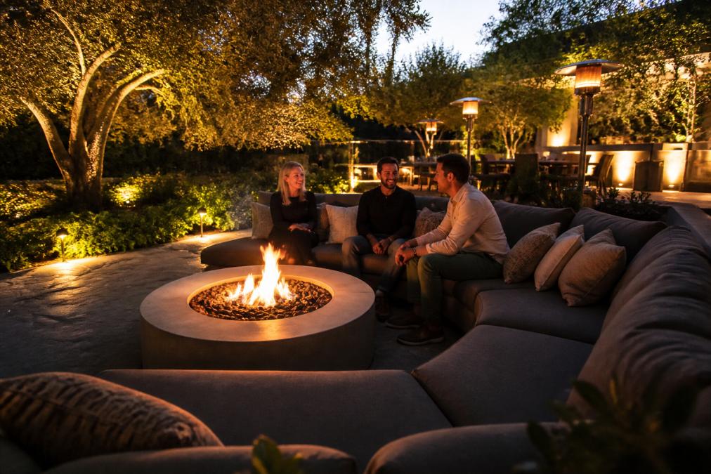 Evening residential backyard with warm amber and white ambient lighting woven through trees, a modern fire pit surrounded by comfortable outdoor sectional seating in the foreground, people gathered around the fire, and softly lit landscaping plants creating depth and texture