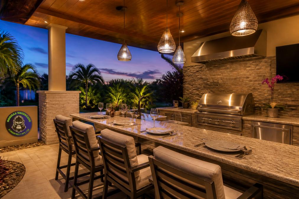 A modern outdoor kitchen with a built-in stainless grill, stone countertops, bar seating with cushioned stools, and warm pendant lighting glowing over the counter at dusk