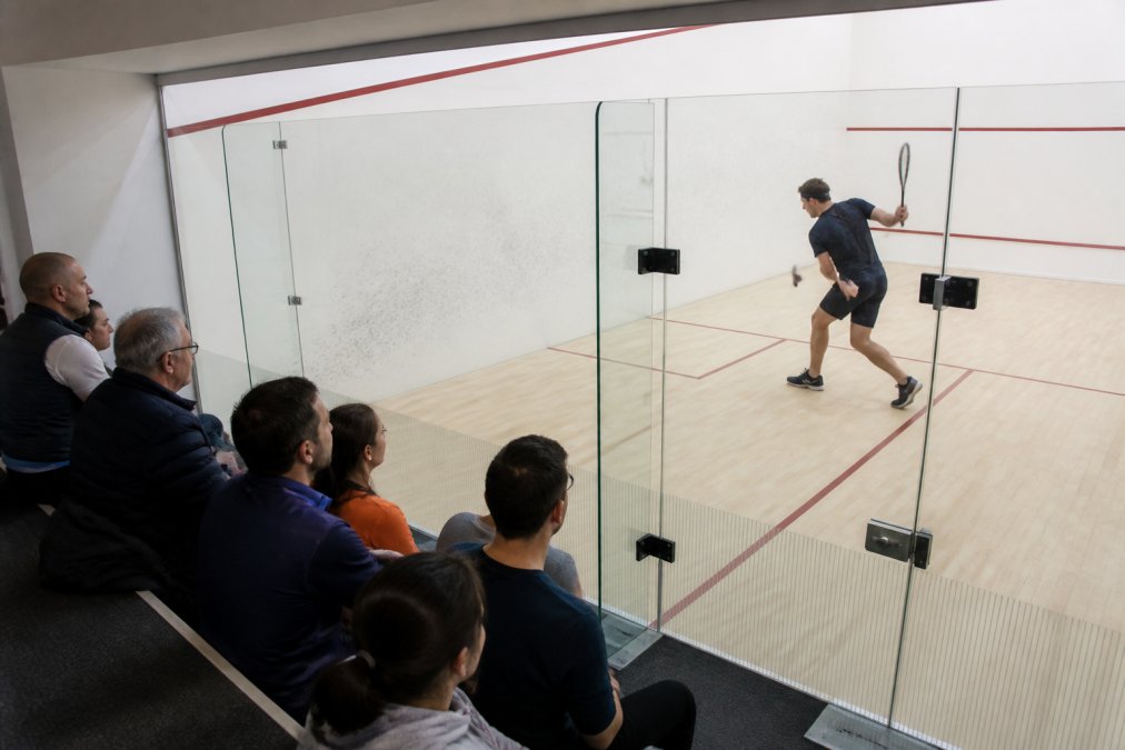 A squash player mid-swing inside a modern glass-backed court, with spectators watching through the transparent wall from a gallery seating area