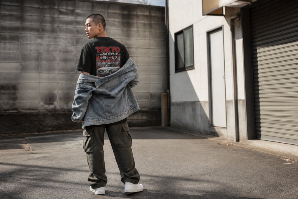 Person wearing an oversized heavyweight graphic tee with a minimal front logo and bold back artwork, layered under an open vintage denim jacket, paired with relaxed cargo pants and minimalist white sneakers, standing in an urban alleyway with natural morning light casting soft shadows