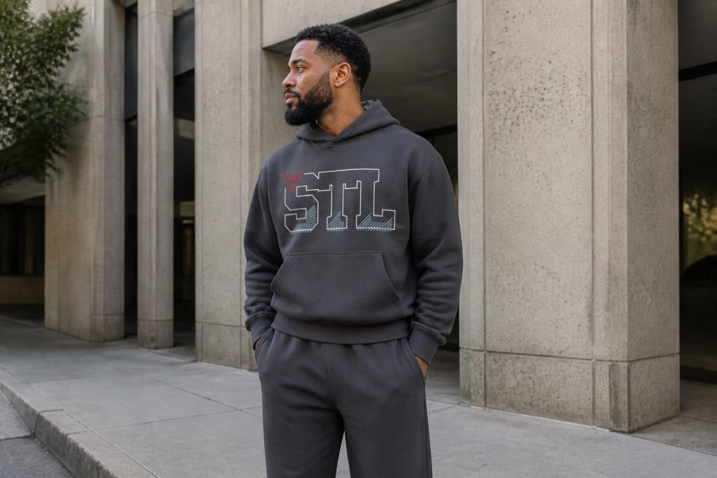 A 30-something man wearing a premium boxy hoodie in charcoal gray and tailored sweatpants, standing confidently in an urban setting with soft natural morning light, showing how elevated basics fit a mature streetwear aesthetic