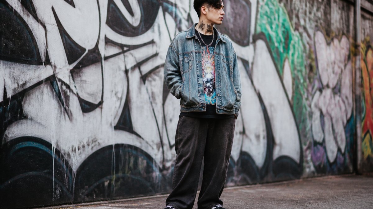 Young adult in a vintage denim jacket and wide-leg trousers posing against graffiti walls for 2026 streetwear trends.