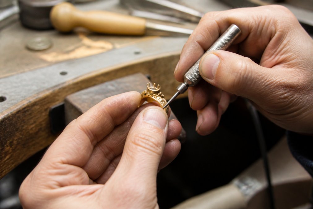 Jeweller's hands working on a delicate gold ring with precision tools in a workshop setting, showing intricate handcrafted detail with natural workshop lighting