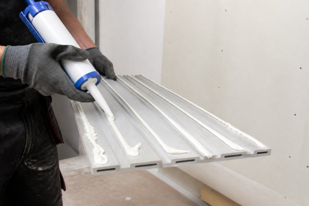 Close-up of hands wearing work gloves applying white construction adhesive to the back of a grey slat wall panel, showing even bead placement across the panel surface against a prepared white wall