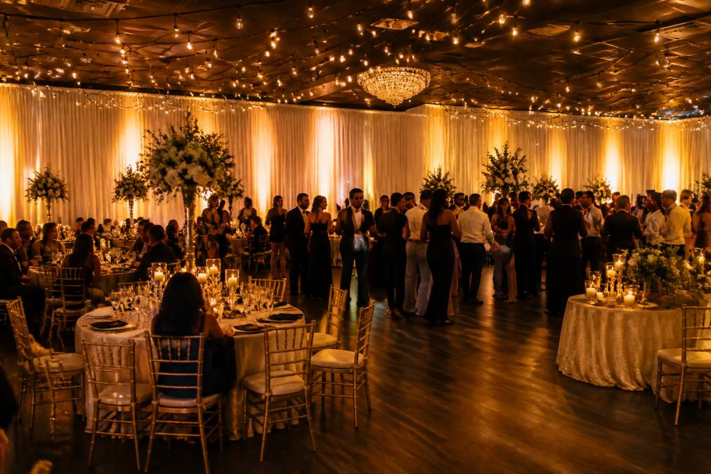 Elegant indoor wedding reception with warm uplighting illuminating draped walls, string lights suspended overhead, and candlelit tables with guests mingling and dancing