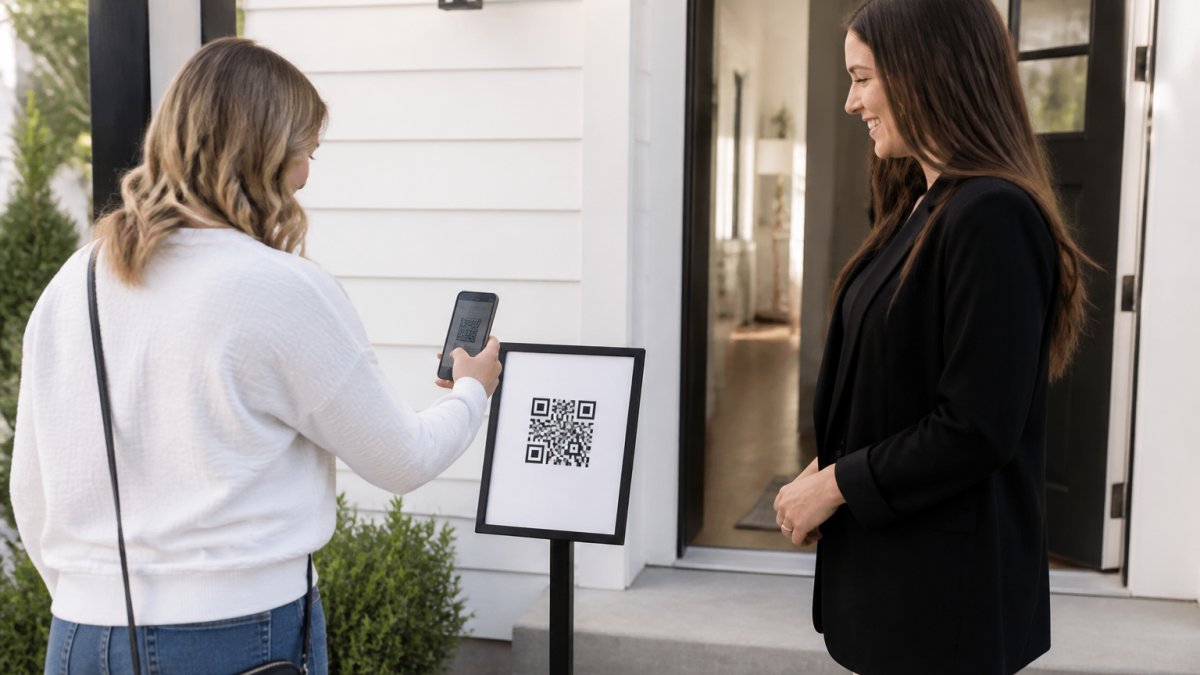 A real estate agent at an open house holding a smartphone showing a QR code sign on a pedestal near the front door, with a visitor scanning it on their own phone in a bright, modern entryway