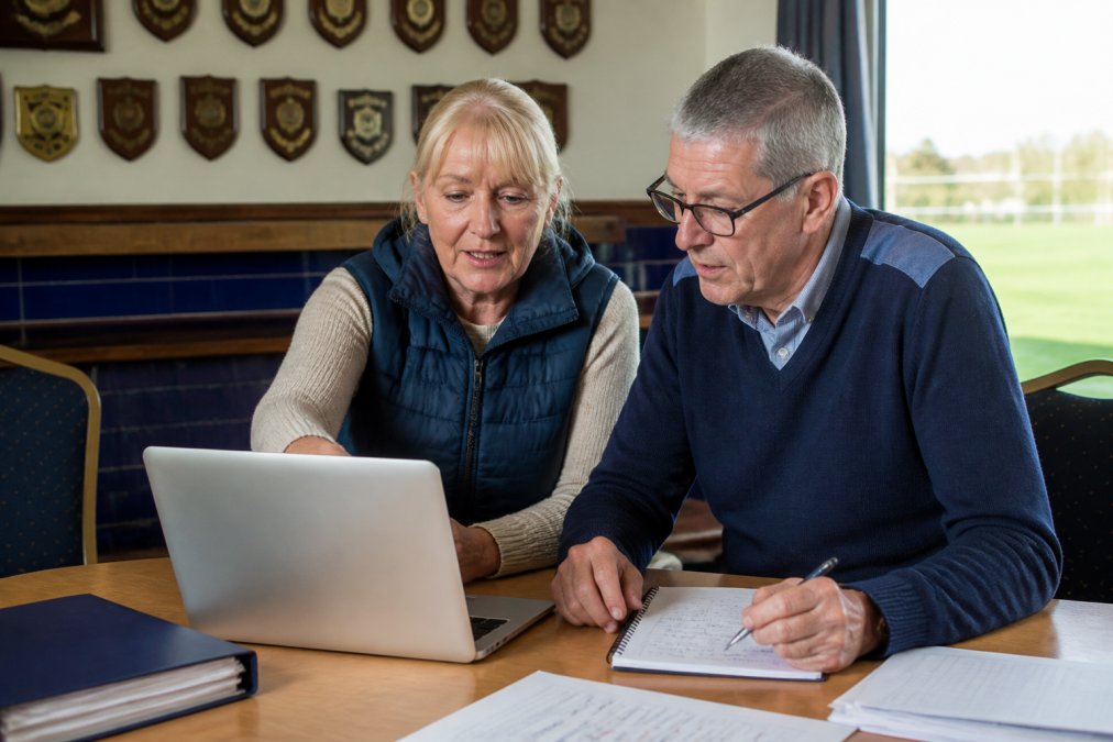 A volunteer club secretary showing a treasurer how to use a laptop at a clubhouse table, papers and a notebook nearby