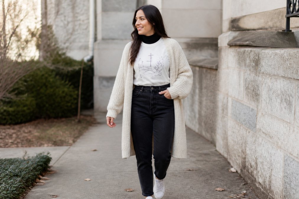 Woman wearing a graphic faith-based t-shirt layered with a fitted black mock-neck undershirt visible at the neckline, topped with an oversized cream cardigan that falls to mid-thigh, paired with high-waisted dark jeans and white sneakers
