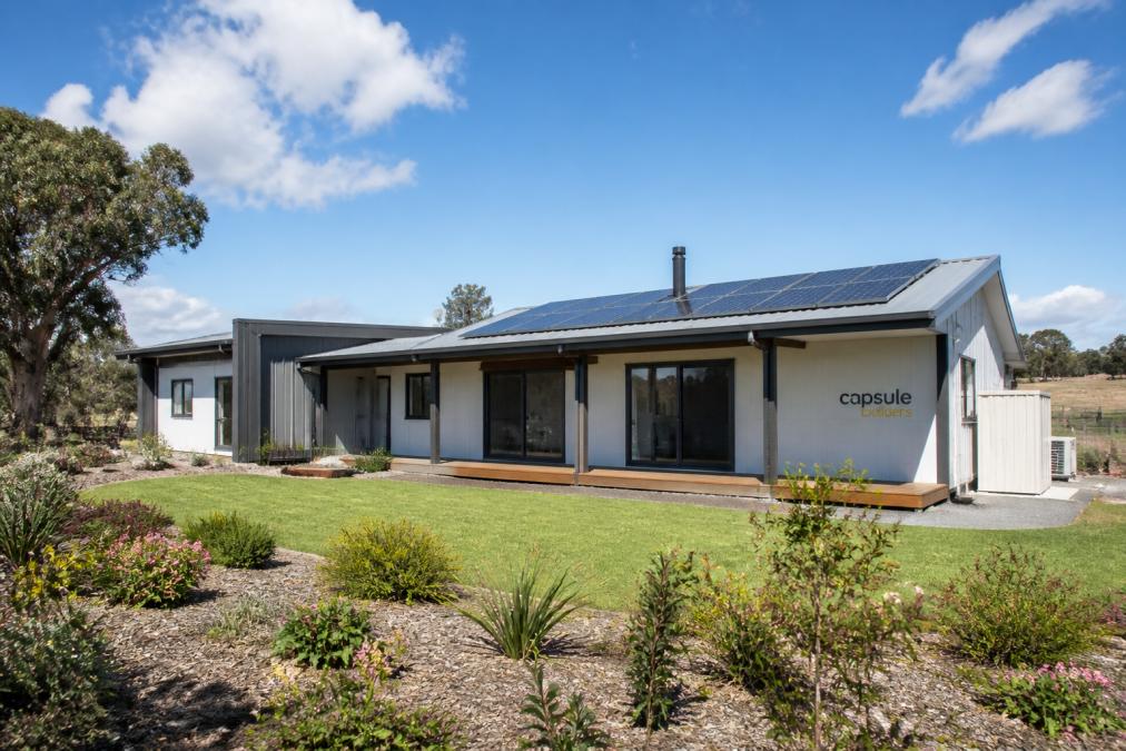 A completed steel-framed modular home on a spacious regional block, with solar panels on the roof and a well-maintained garden, photographed under bright midday sunlight with clear blue sky
