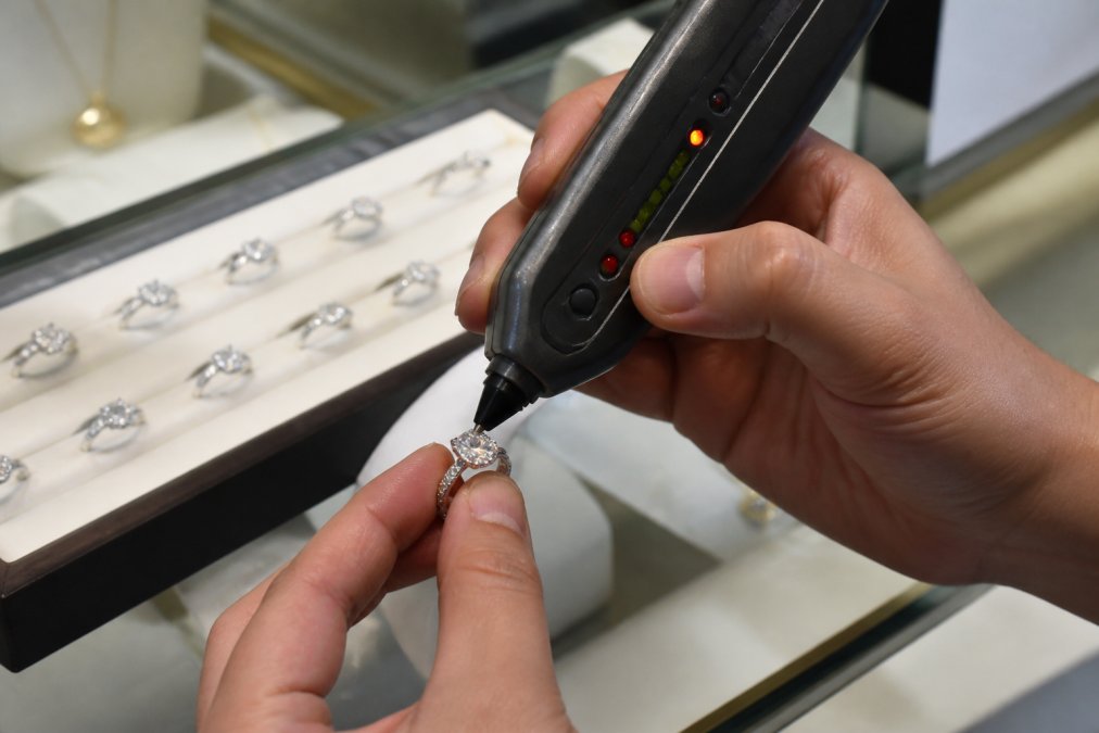 Close-up of a hand pressing a handheld diamond tester pen against a sparkling moissanite ring on a well-lit jewelry counter
