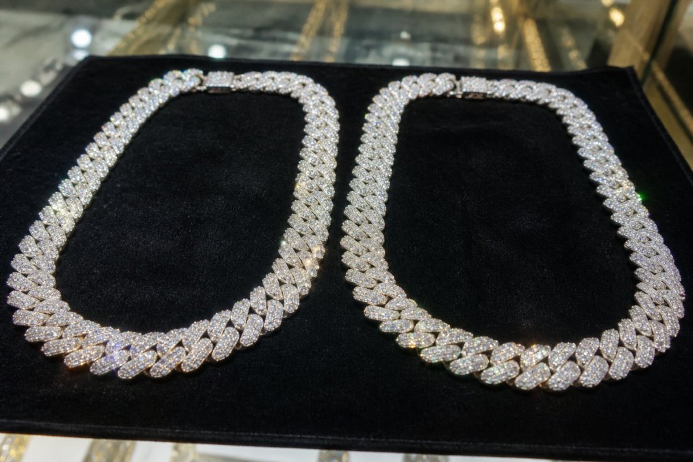 Two iced-out Cuban link chains resting side by side on dark velvet under bright jewelry-store lighting, one VVS1 moissanite and one diamond, both throwing visible rainbow fire and white sparkle across the fabric
