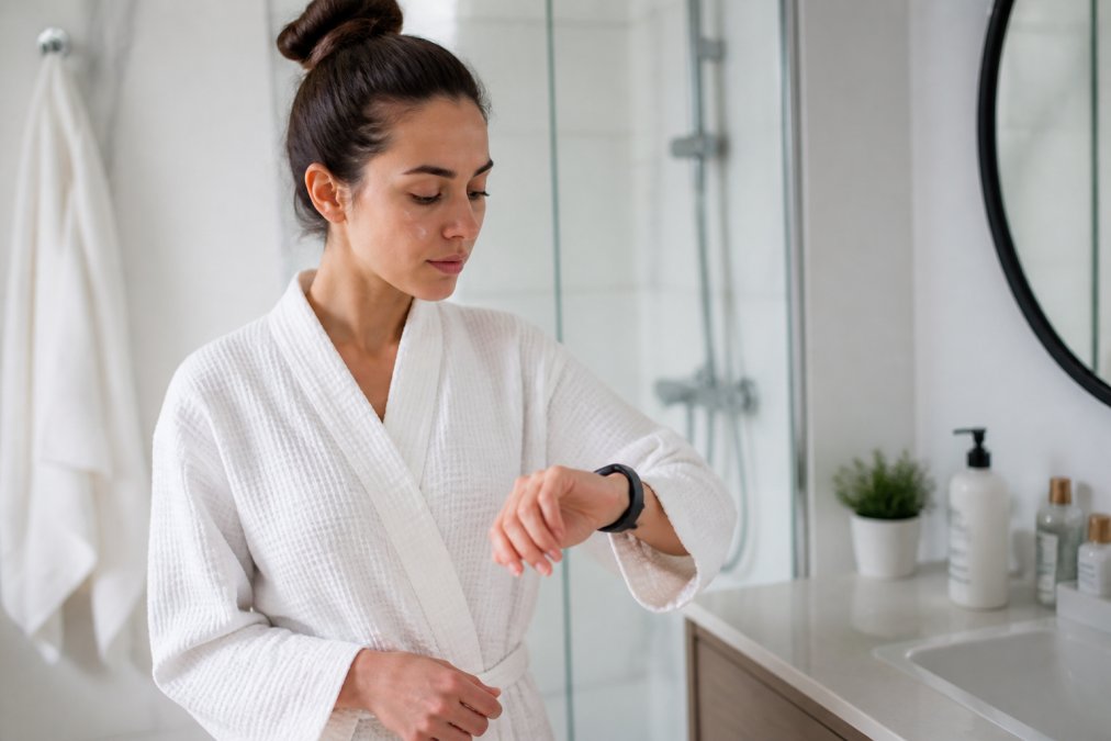 Person überprüft die Zeit auf ihrer Smartwatch, während sie zwischen Hautpflegeanwendungen in einem hellen Badezimmer wartet, was die Bedeutung von Geduld in einer Morgenroutine demonstriert