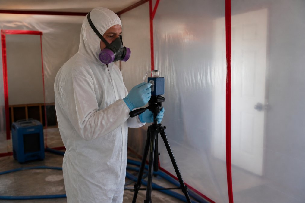 Environmental professional in protective gear collecting air samples with specialized equipment in a remediation work zone with containment barriers visible