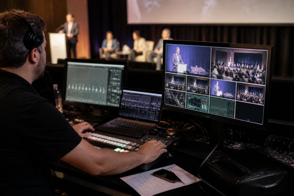 Professional technician at on-site control station with multiple monitors displaying camera feeds, audio levels, and streaming metrics during a corporate event broadcast
