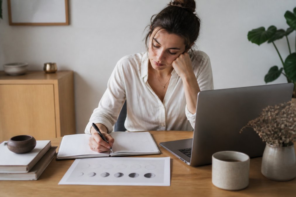 Berufstätige Frau an ihrem minimalistischen Schreibtisch mit Mondkalender neben ihrem Laptop, notiert ihre Wochenplanung und schaut auf den Mondzyklus