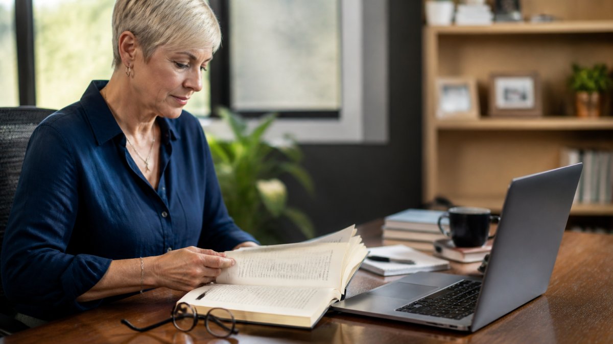 A consultant reviewing a printed book alongside a laptop showing an email list and client pipeline in a bright office