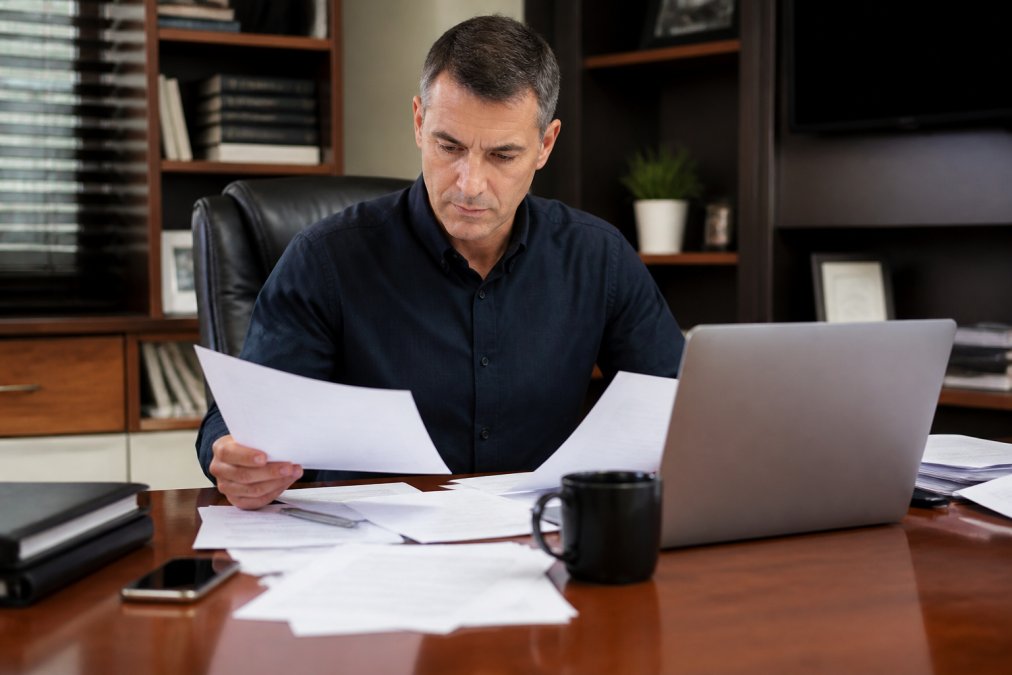 Professional business owner reviewing documents at desk with laptop, analyzing business credit information with focused attention and organized workspace