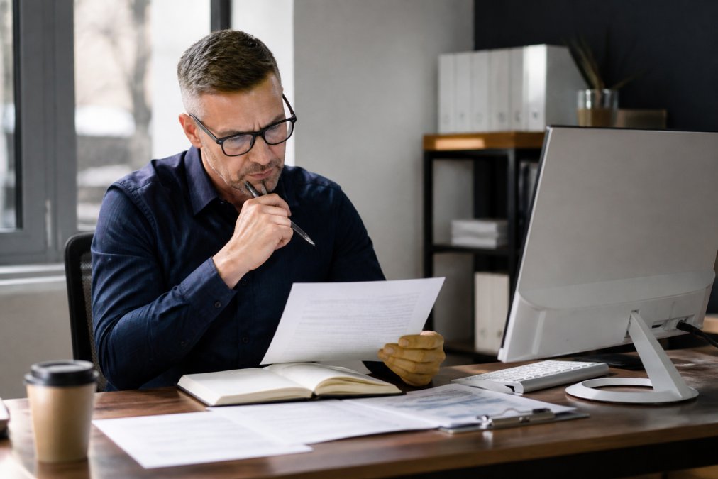 Professional business consultant sitting at a desk reviewing equipment brokerage contracts and credentials on a computer screen, with a notepad and pen for notes, in a modern office with natural window lighting