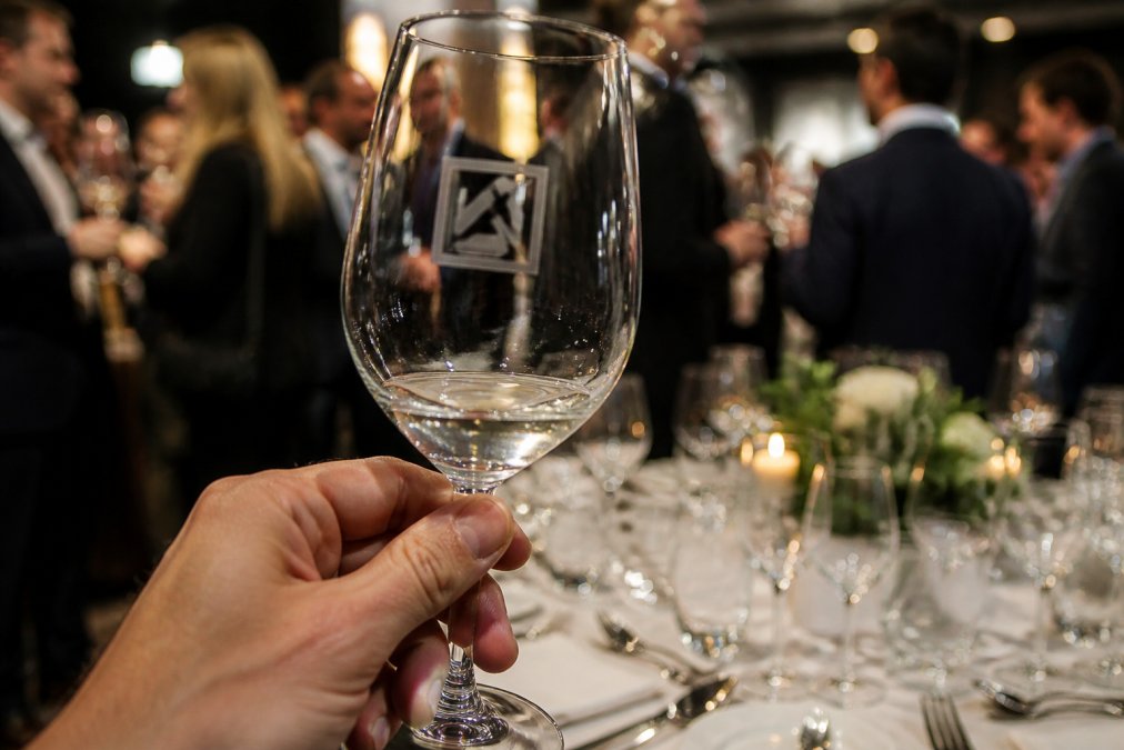 Close-up of an elegantly engraved wine glass with a corporate logo, held at a business event with guests and table settings in soft focus behind it