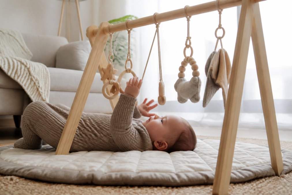 An infant lying on their back during tummy time beneath a wooden activity gym with natural hanging toys, reaching upward with focused concentration toward wooden rings and fabric elements