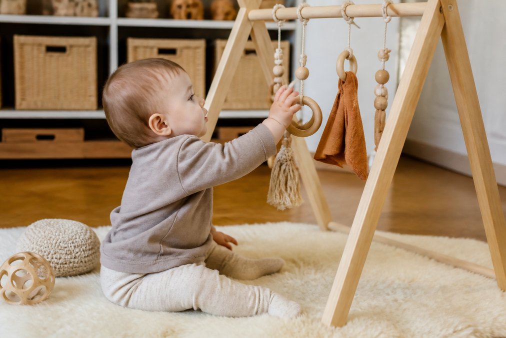 A toddler approximately one year old sitting near a wooden activity gym, reaching toward and grasping wooden rings and natural fabric elements with deliberate hand movements and focused attention