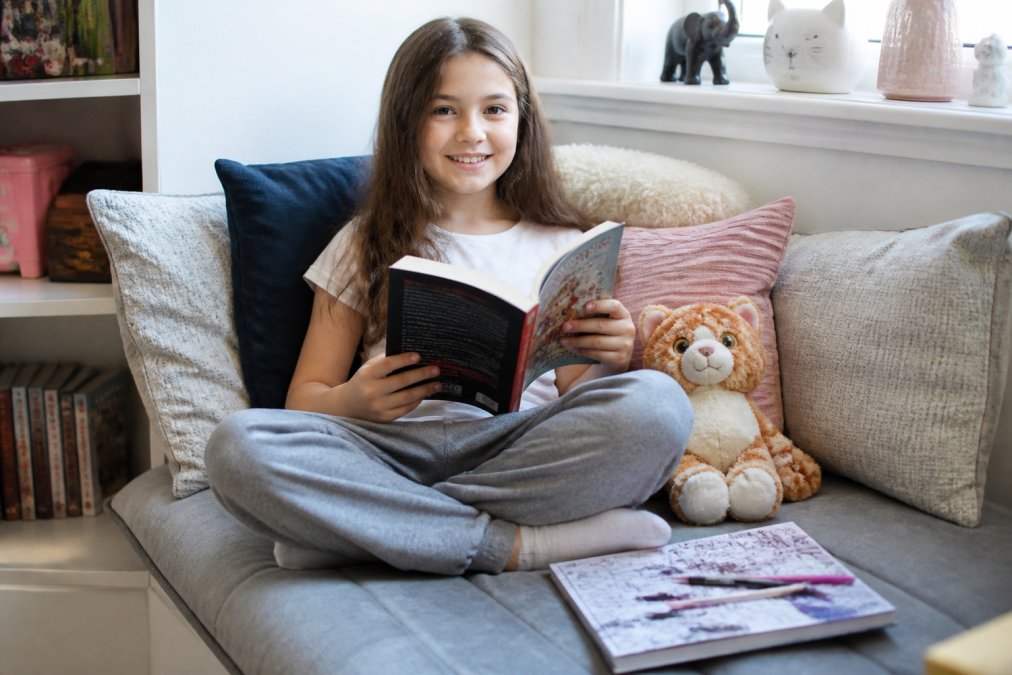 A 9-year-old child sitting cross-legged on a cozy reading nook, smiling while holding a softcover book from the Miso & Kili series, with a plush cat toy and activity book nearby
