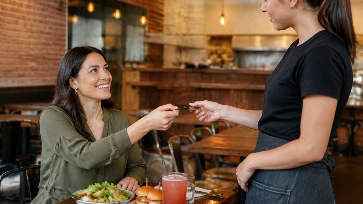 Diner smiling while showing their membership card or app to a restaurant server at a local eatery, with food and drinks on the table, warm lighting
