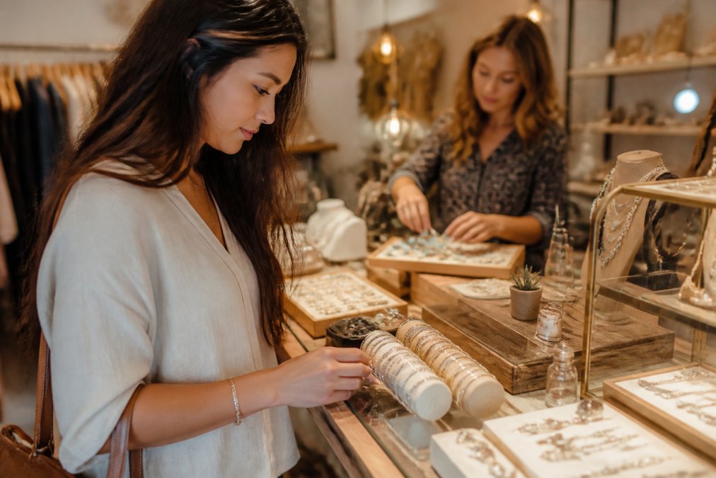 Woman browsing handcrafted sterling silver bracelets in a boutique as a shopkeeper arranges gemstone jewelry
