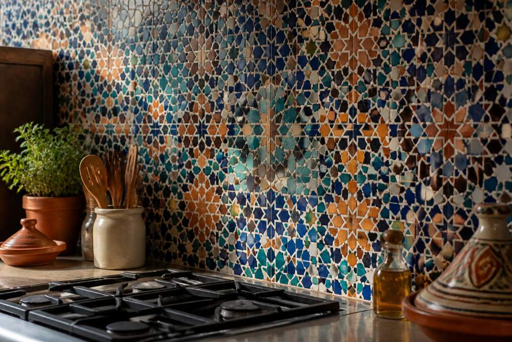 Close-up of intricate handmade Moroccan zellige tiles with geometric patterns in rich blues, terracotta, and earth tones installed in a kitchen backsplash with natural light highlighting the dimensional glaze