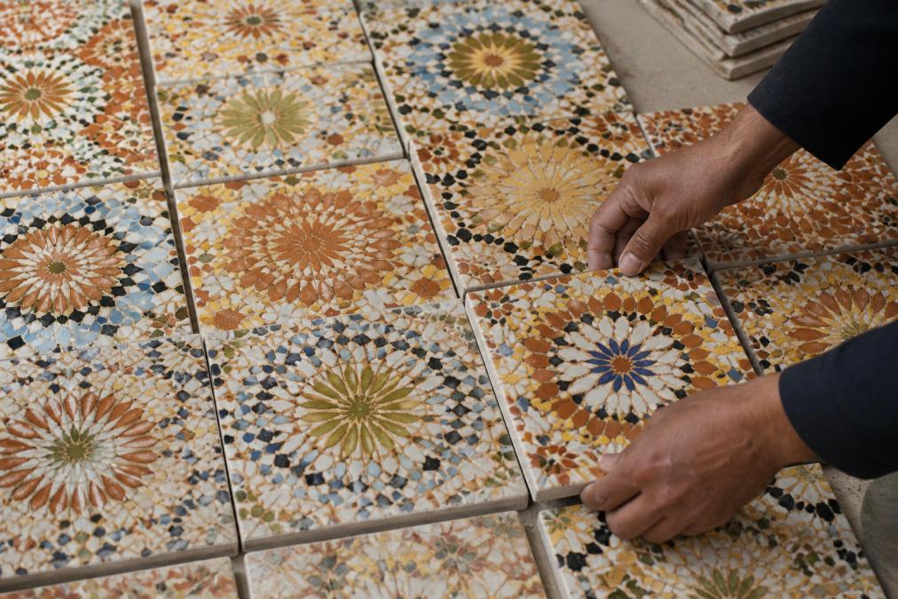 Close-up of handcrafted Moroccan tiles laid out in a grid pattern showing natural color variations ranging from deep terracotta to light cream tones, with hands arranging tiles to blend colors, natural workshop lighting