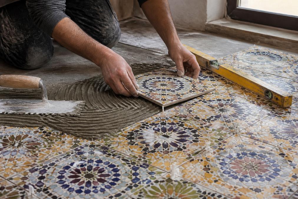Professional installer using a tile spacer and level on Moroccan floor tiles, showing proper trowel technique with ridged thin-set mortar, hands positioning a handcrafted tile with leveling shims visible, natural daylight from workshop window