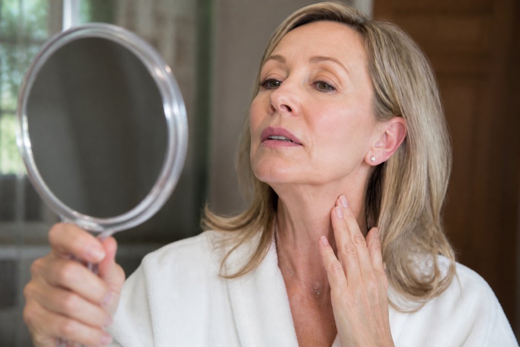 A close-up photograph of a woman in her 40s examining her jawline and neck in a handheld mirror, with soft natural lighting highlighting smoother skin texture and improved contour