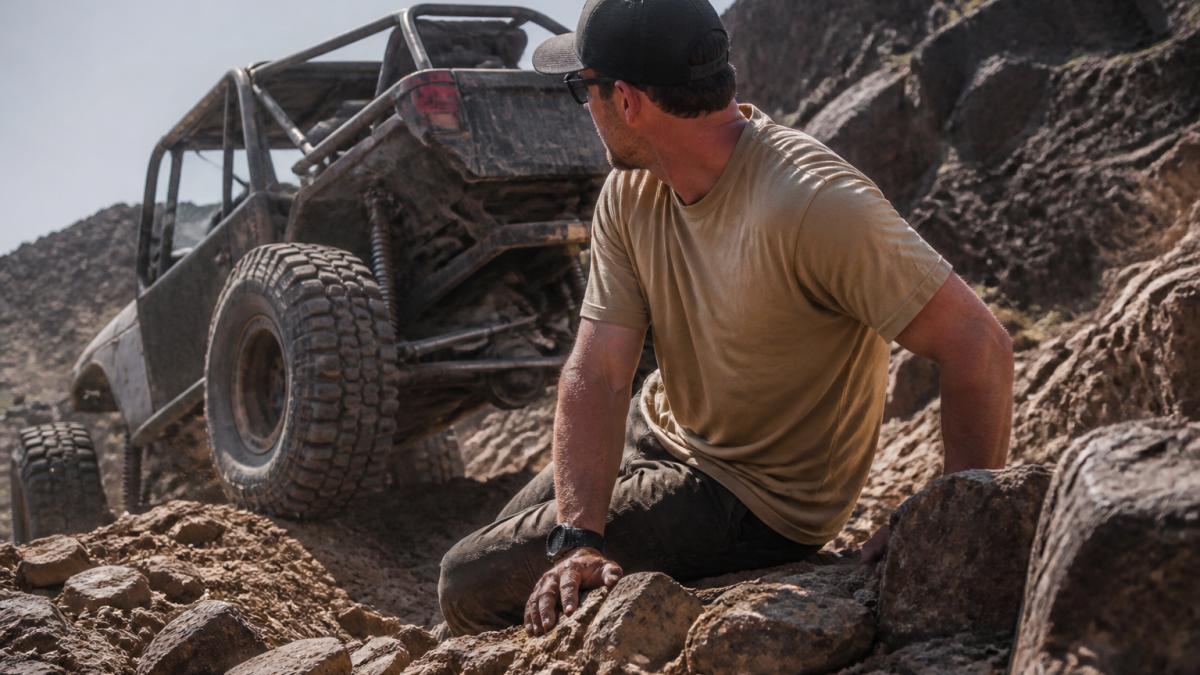 Off-road enthusiast wearing premium heavyweight tee during rock crawling on rocky terrain, with visible dust and natural outdoor lighting, showing the garment holding up during active trail use