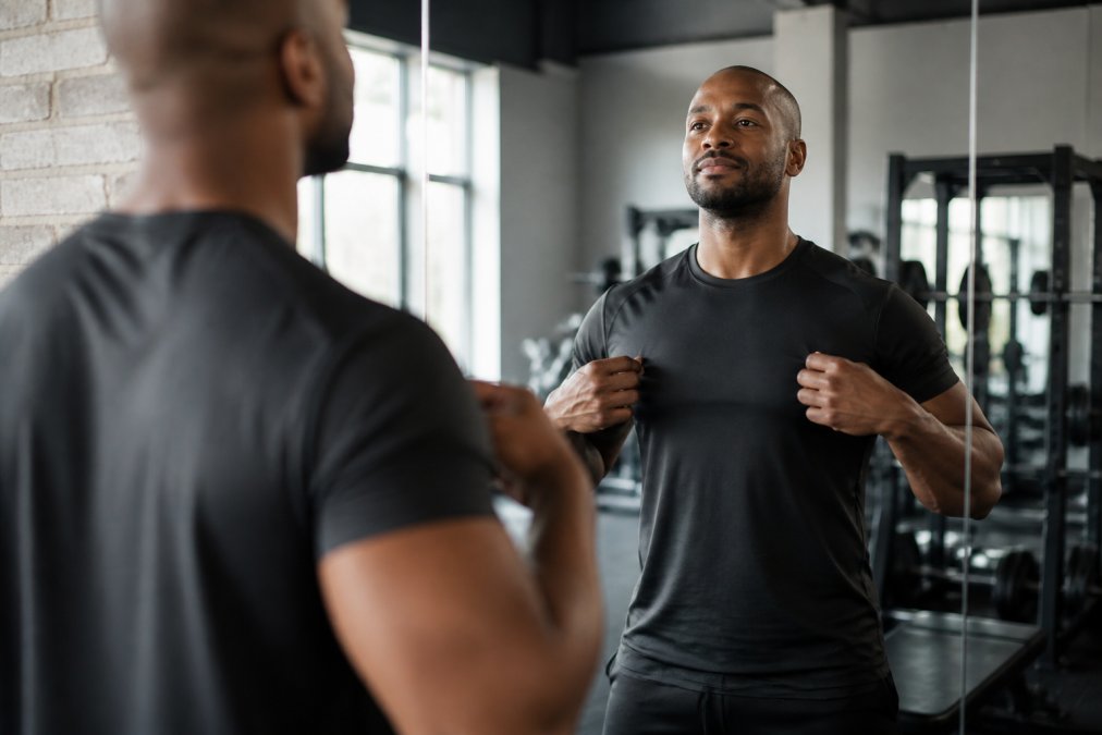 A person in a gym looking at their reflection in a mirror, adjusting a motivational t-shirt, with a confident and determined expression. Soft natural light, clean modern gym background.