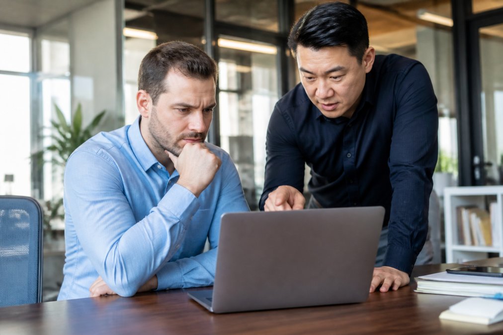 A small business owner in a modern office looking at a laptop with a concerned expression, while an IT professional points at the screen explaining a solution