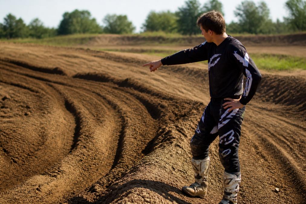 Motocross rider walking the track, examining deep ruts and berms in the dirt, pointing at terrain features while studying the racing line with focused intensity