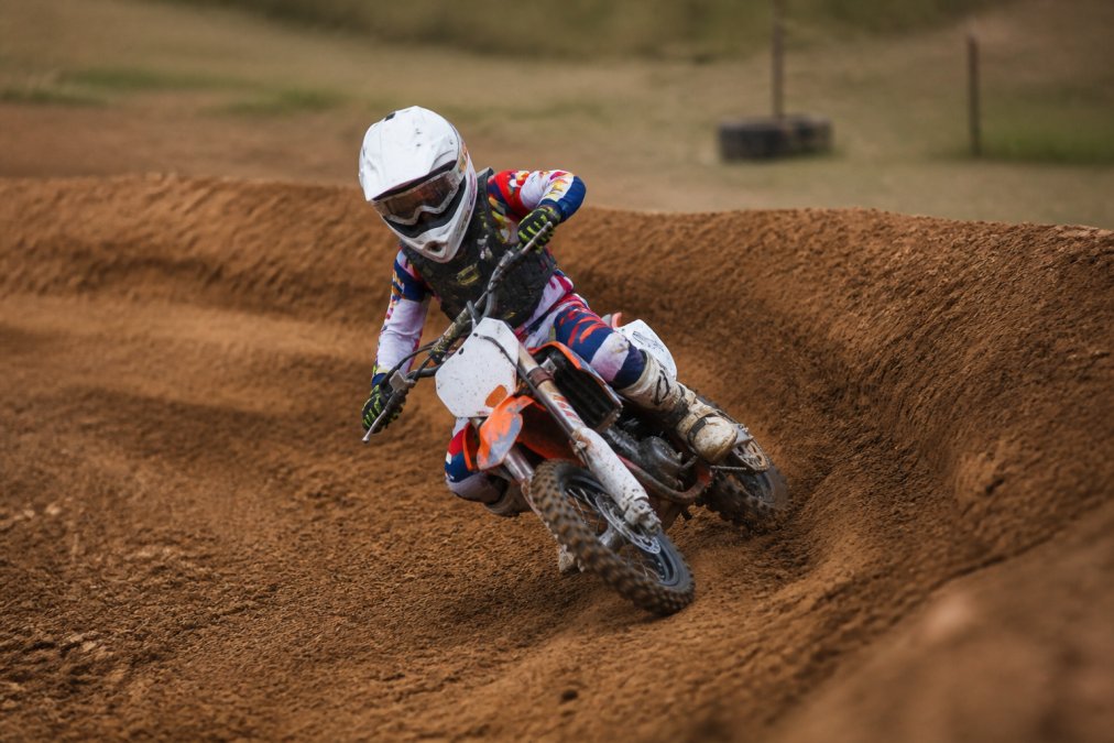 Young motocross rider leaning into a berm on a dirt bike, demonstrating proper inside line technique with body positioned off the seat, throttle hand steady, bike angled at the apex of the turn
