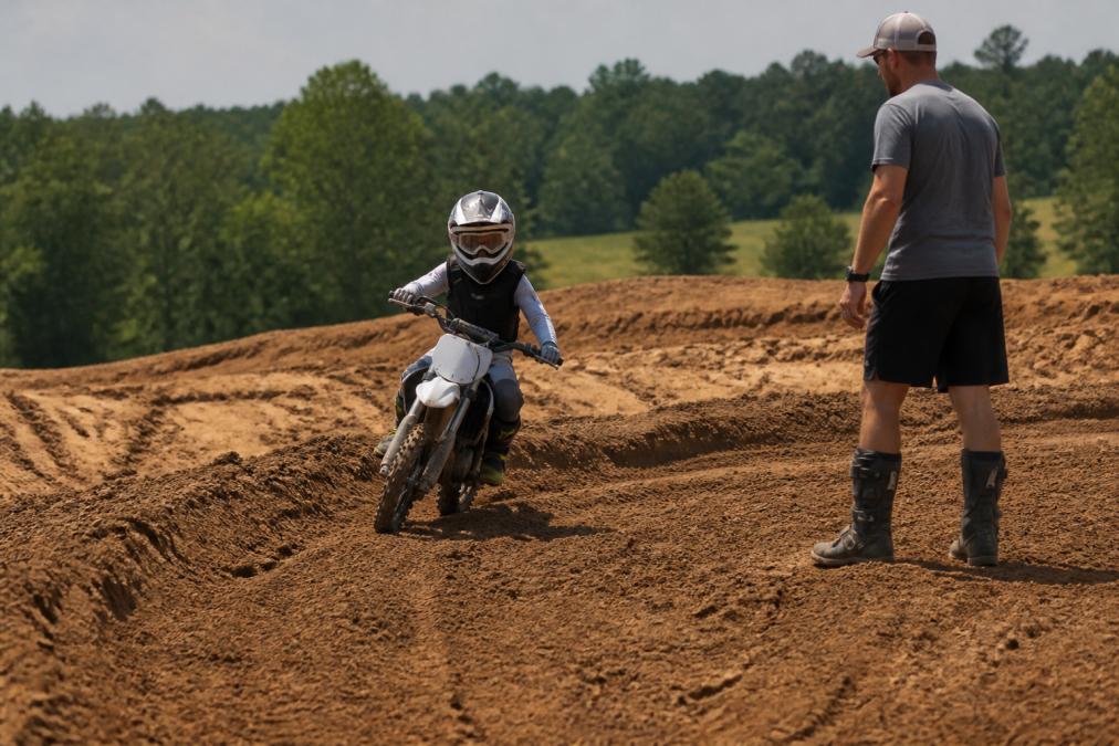 A young rider on a small dirt bike navigating a prepped sandy loam motocross track with a coach standing nearby, trees and countryside in the background