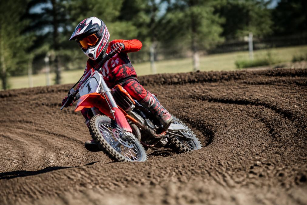 Young rider on a compact motocross bike with aggressive fork angle and tight geometry, navigating a banked turn on a closed-course track with natural daylight
