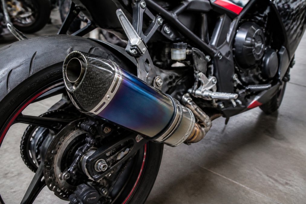 Close-up van een glanzende titanium Yoshimura slip-on uitlaat op een sportmotor, met zichtbare constructiedetails en finish, zonlicht weerkaatsend op het oppervlak