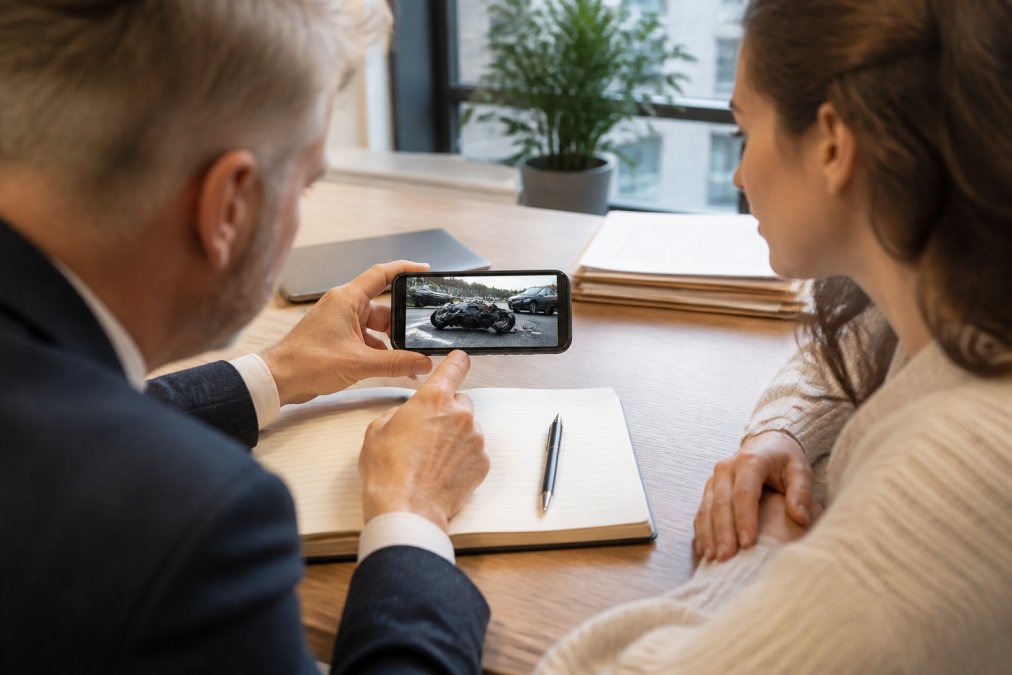 Hands holding a smartphone displaying dashcam footage of a road traffic accident, with a notebook and pen on desk nearby for documenting claim details, natural office lighting