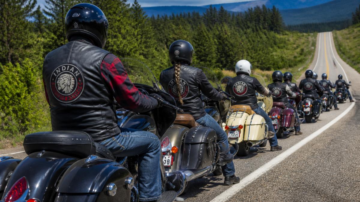 Group of motorcyclists lined up on a scenic road during a charity ride, wearing vests and helmets with trees and open road stretching behind them