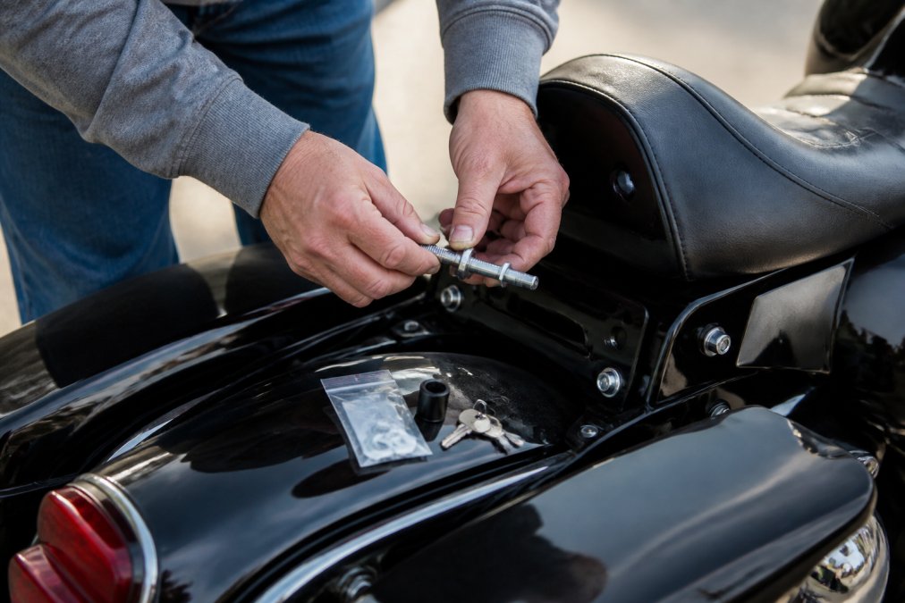 A rider's hands threading a locking seat bolt into a Harley-Davidson touring motorcycle seat mount, with a small hardware kit and key visible on the bike's fender in natural outdoor light