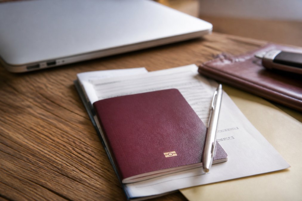 Close-up van documenten, een paspoort en een pen op een bureau met een laptop ernaast, wat de voorbereiding op import symboliseert