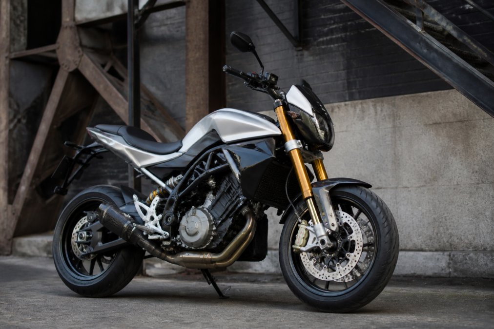 Husqvarna NUDA motorfiets in glanzend zilver geparkeerd van opzij, met focus op de voorkant en de Öhlins-vering duidelijk zichtbaar, tegen een industriële achtergrond