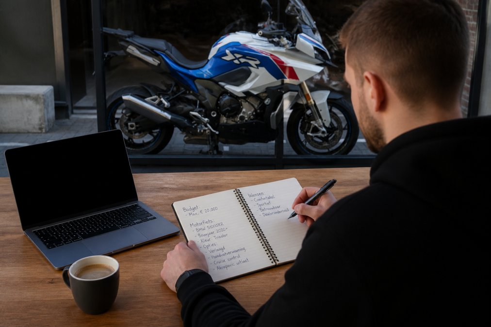 Person writing detailed motorcycle specifications and budget notes in a notebook at a desk with laptop and coffee, natural office lighting