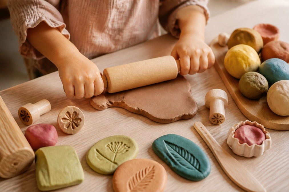 Kinderhanden boetseren figuren met natuurlijke klei en houten rollers op een houten tafel.