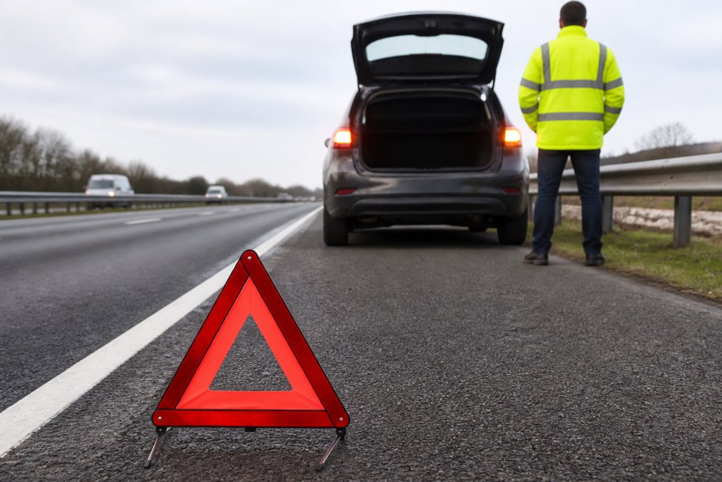 Motorway Breakdown Rules: Highway Code Guide Driver standing safely behind their stationary vehicle on a motorway hard shoulder wearing high-visibility yellow jacket, with hazard warning lights activated and a warning triangle positioned on the road surface behind the vehicle