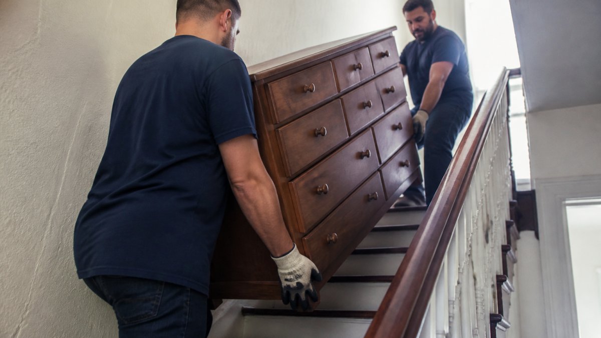 Two movers in work gloves carrying a large dresser up a narrow apartment stairwell, one at the top step guiding the load while the other supports the base, natural light from a hallway window